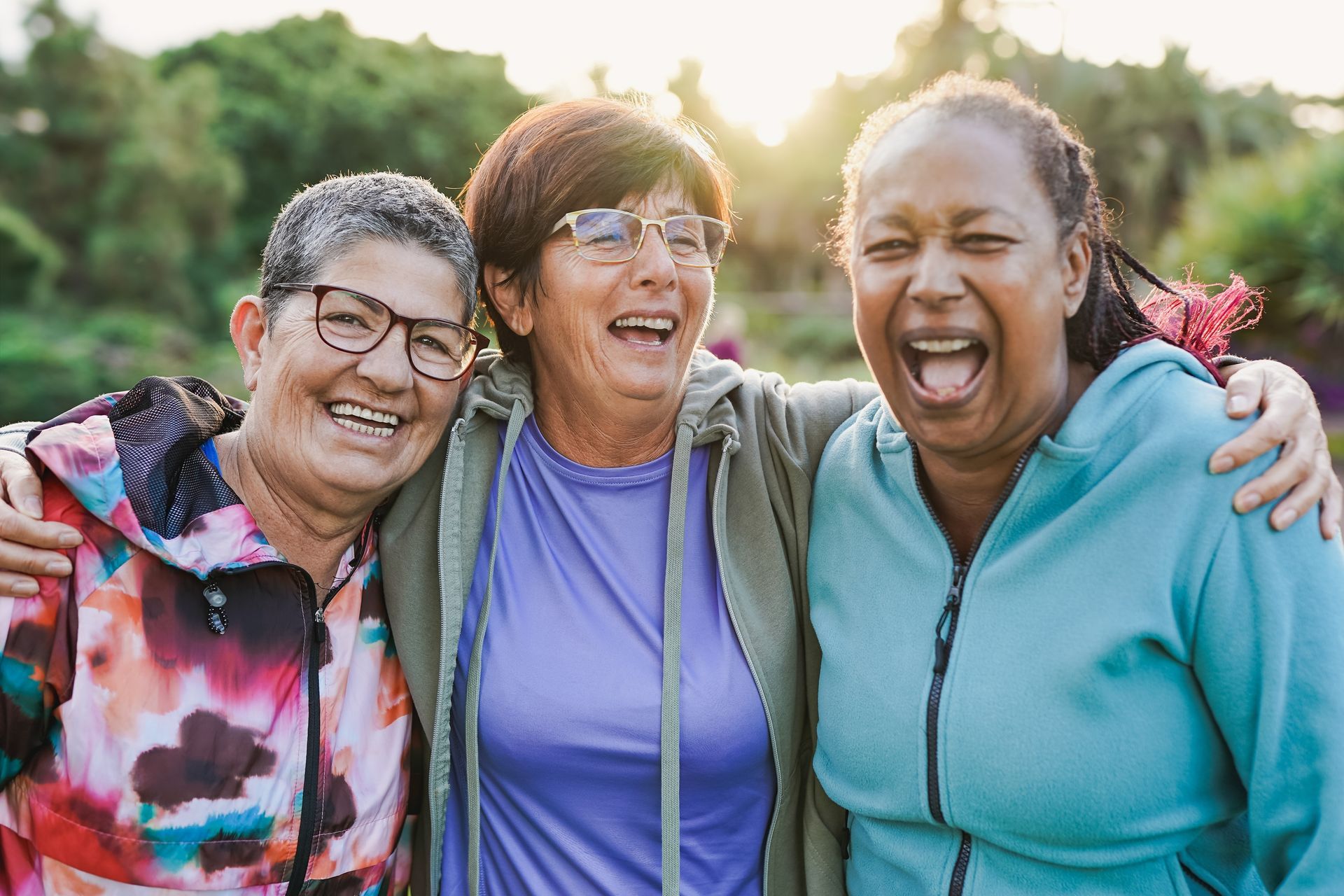 Three diverse women laughing, embracing outdoors. Sunshine, colorful clothing, park setting.