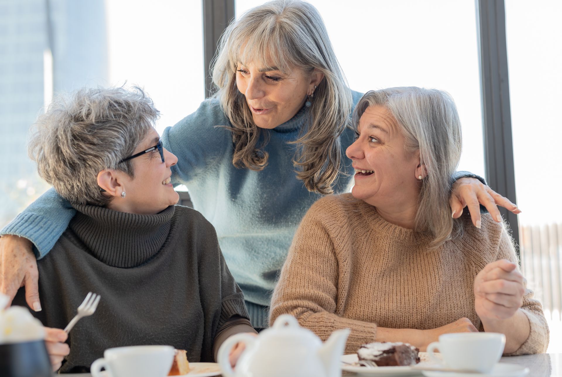 Three older women with gray hair, smiling, talking, enjoying tea and cake at a table.