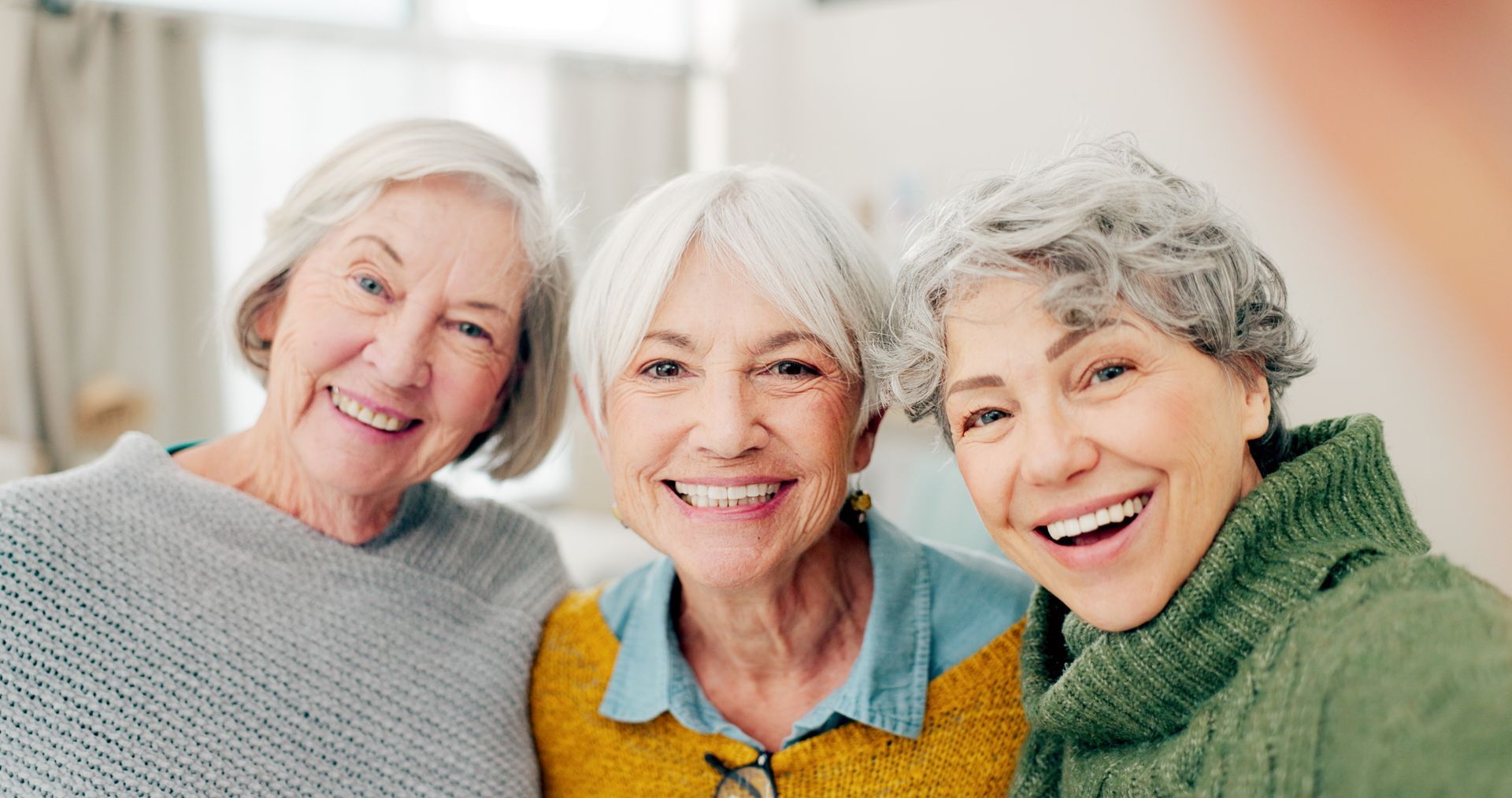 Three smiling older women taking a selfie indoors. They are of different complexions, with gray hair and wearing sweaters.