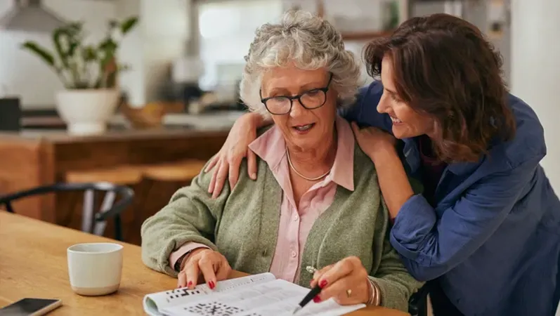 Woman helping another with a crossword puzzle at a table indoors.