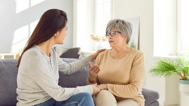 Young woman comforts older woman, touching her arm and hand, both seated on a sofa.