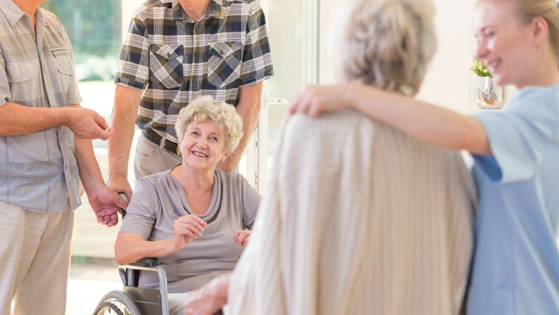 Elderly woman in wheelchair smiles, surrounded by a group of people, with a caregiver's arm around her.