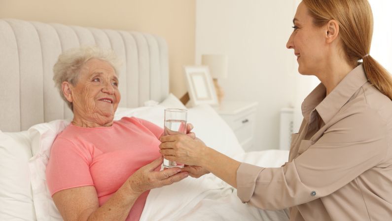 Woman offers a glass of water to an elderly person resting in bed. White sheets, neutral tones.