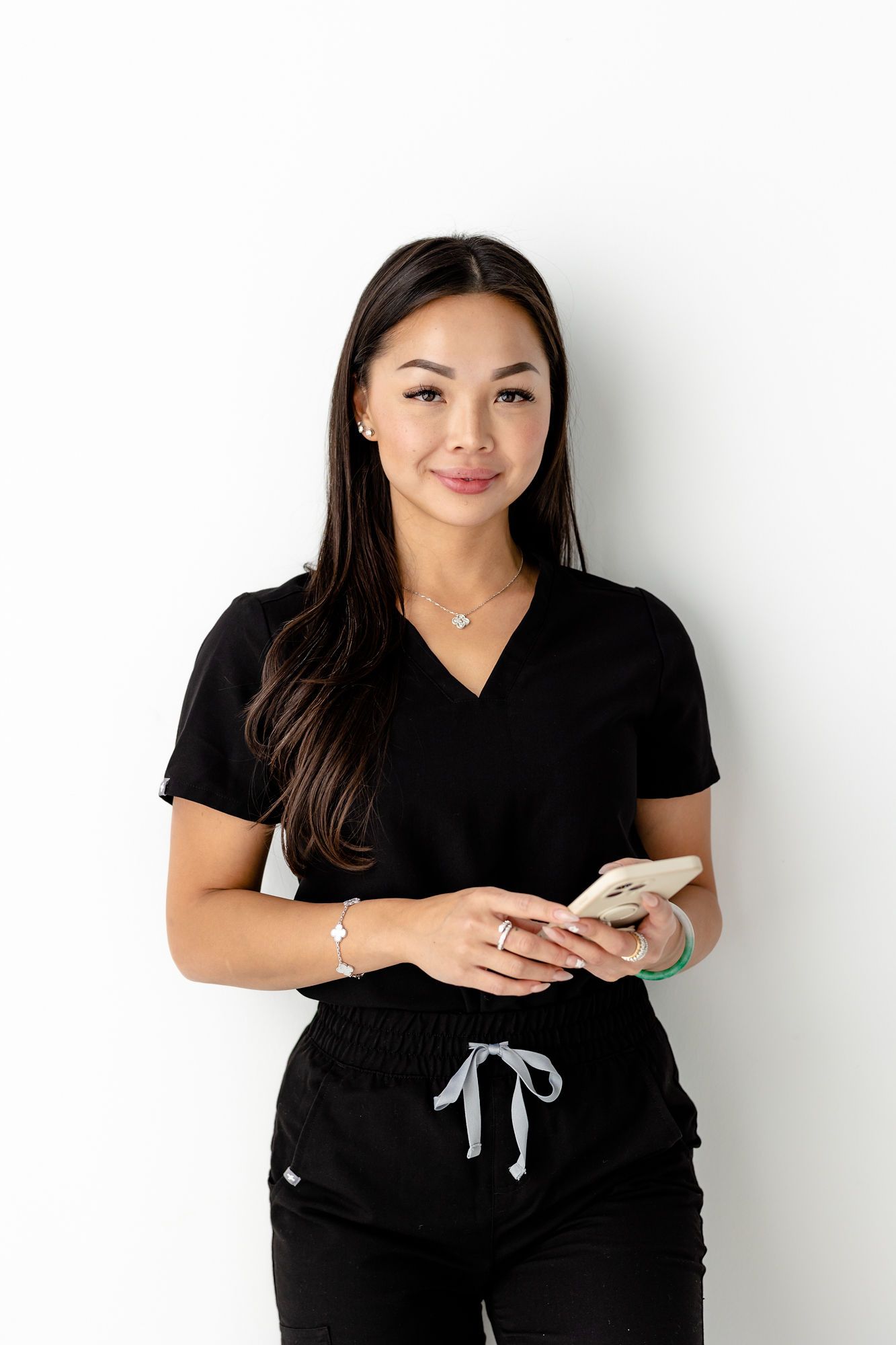 Woman in black scrubs holding a phone, leaning against a white wall.