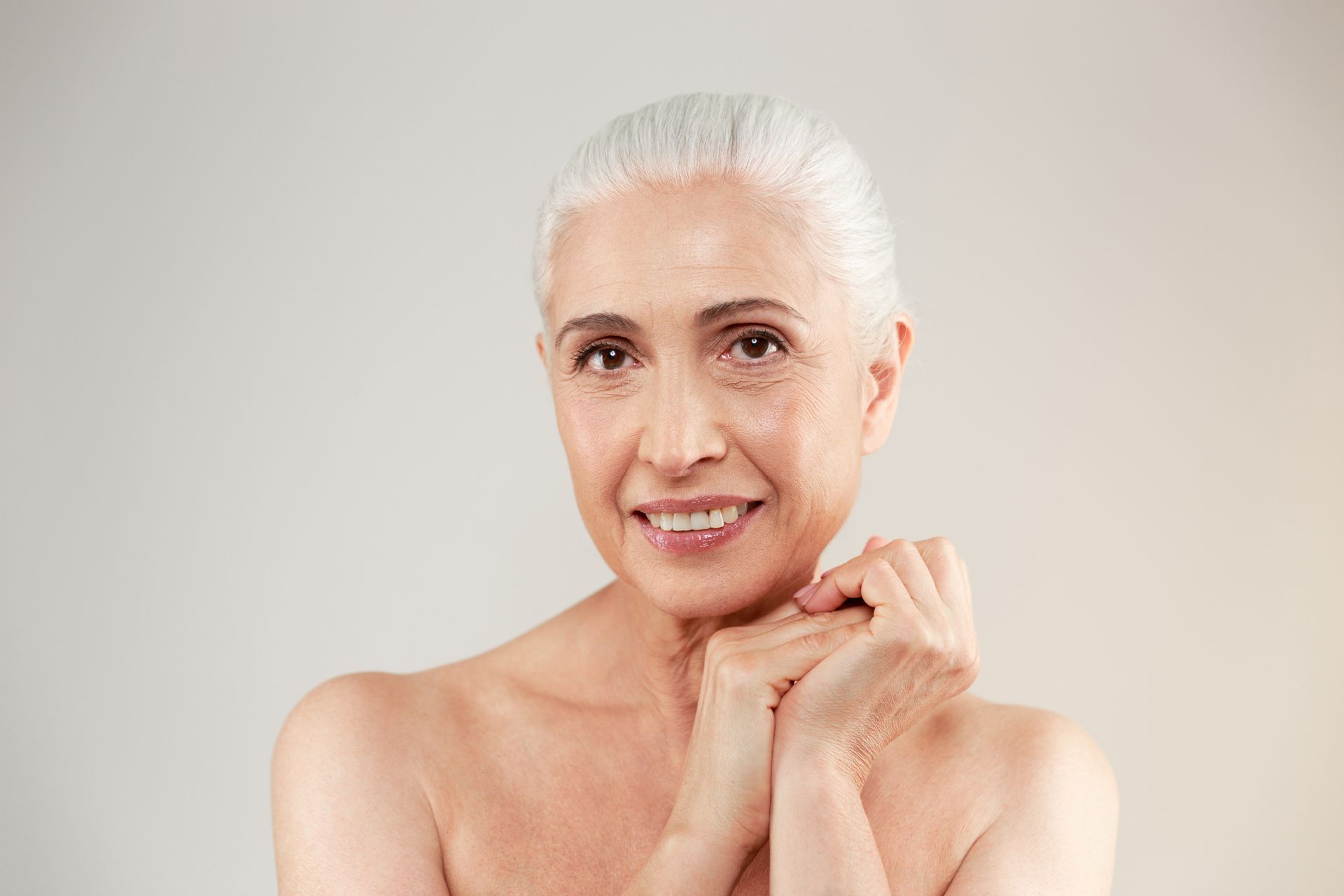 Woman with short white hair, smiling, hands clasped, bare shoulders, soft lighting.