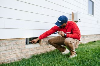 A man in a red shirt and blue hat is kneeling down in front of a house.