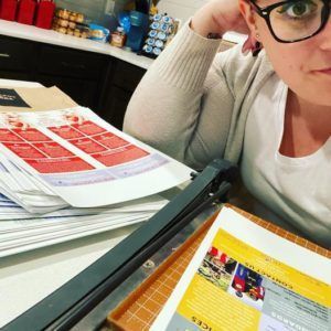 Person with glasses leans on arm, looking at paperwork with a paper cutter. Kitchen counter setting.