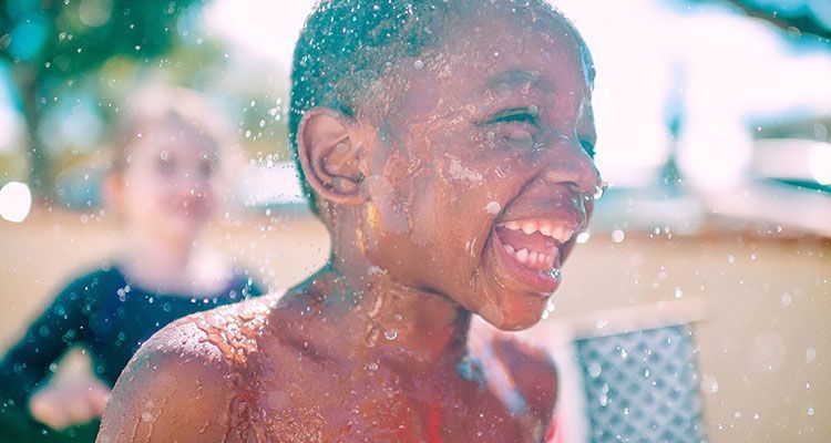 Boy laughing, getting sprayed with water; another child in background.
