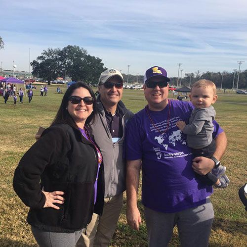 Four people pose outside on a sunny day. One holds a baby. People are walking in the background.