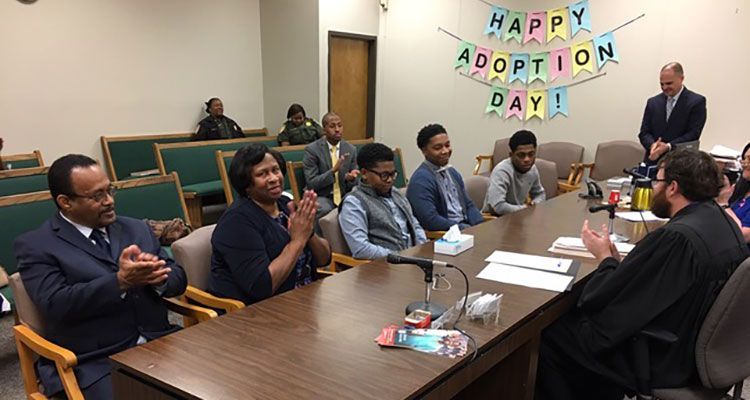 A family celebrates an adoption in a courtroom with a judge, a banner, and supportive attendees.