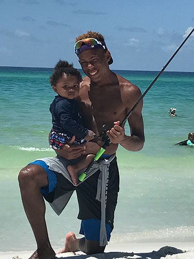 Man kneels on a beach holding a child and a fishing rod. The water is turquoise.