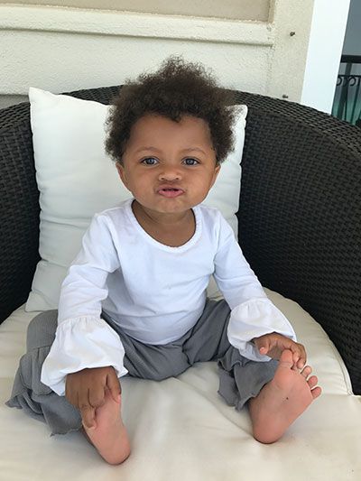 Child with curly hair puckering lips, seated on a woven chair, wearing white top and gray pants.