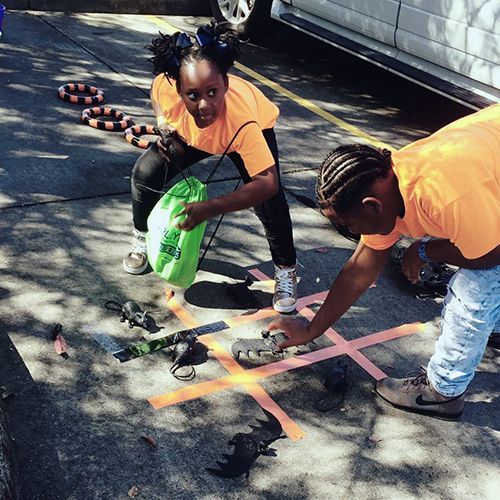 Two children playing tic-tac-toe on pavement, orange shirts, dark hair, bats, rings in background.