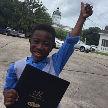 Young person holding a folder with raised arm, thumbs-up gesture, in front of a courthouse.