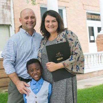Family of three stands in front of a courthouse; woman holds documents, all smiling.