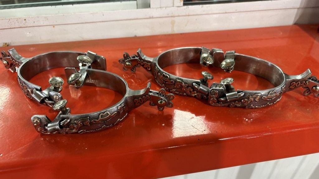 Pair of ornate silver spurs with decorative floral and figure details, resting on a glossy red surface.