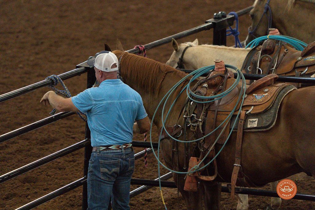Man in blue shirt adjusting rope on a saddled horse in a rodeo arena.