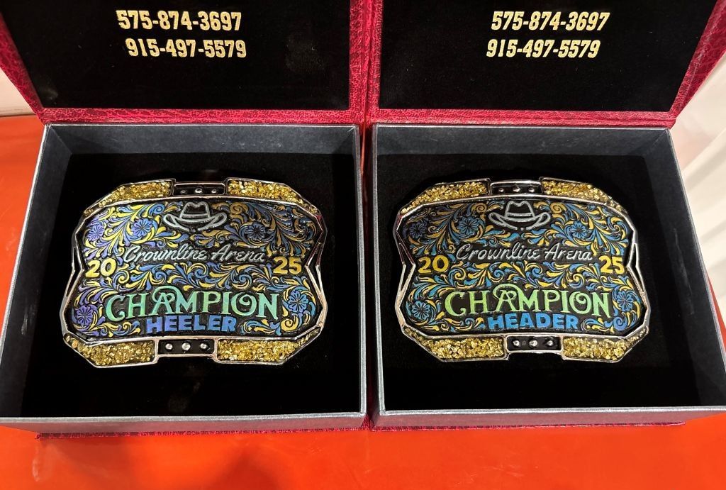 Two Champion rodeo belt buckles in presentation boxes with phone numbers on the lid. The buckles are blue, gold, and silver.