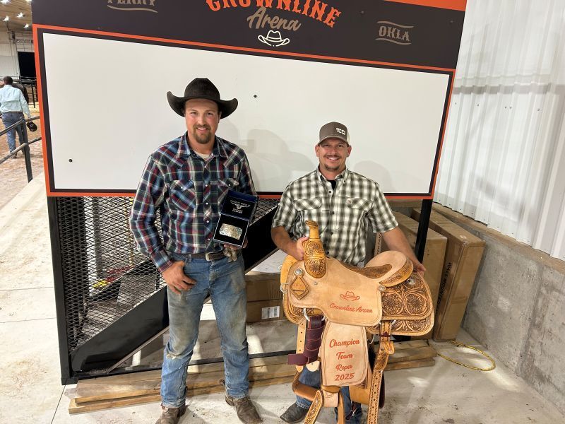 Two men posing with a trophy and a saddle at an arena. One wears a cowboy hat.