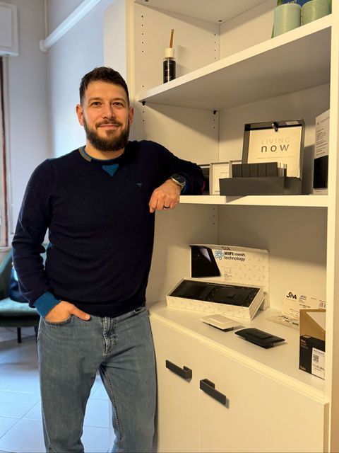 Un uomo con maglione blu e jeans è appoggiato a uno scaffale bianco su cui sono esposti i prodotti. Ha la barba e sorride.