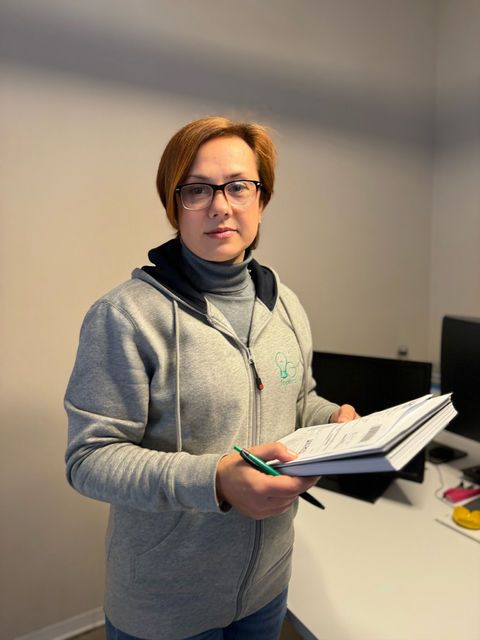 Donna con felpa grigia e occhiali, che tiene in mano un quaderno e una penna in un ufficio.