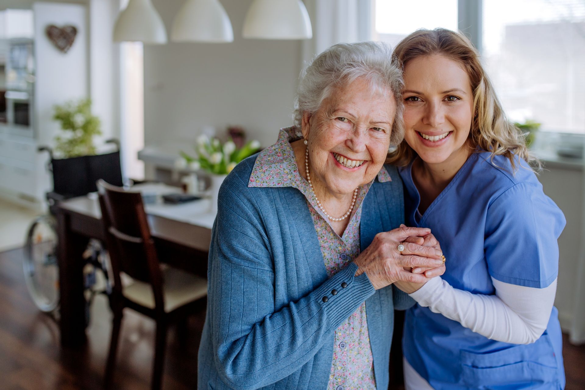 Smiling caregiver in blue scrubs embracing an elderly woman in a bright home dining room