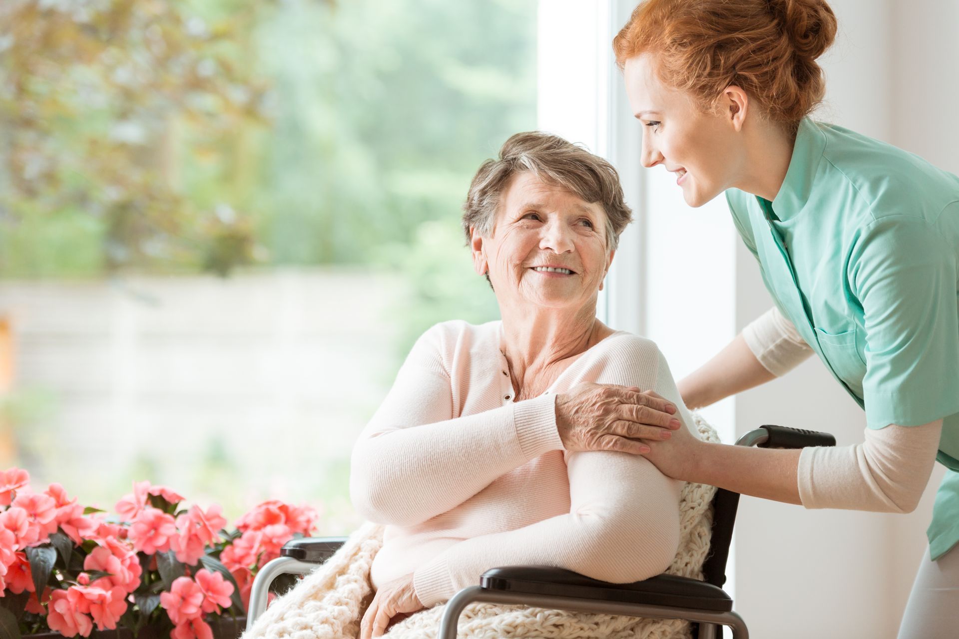 Caregiver helping an elderly woman sit in a wheelchair, with flowers in the foreground.