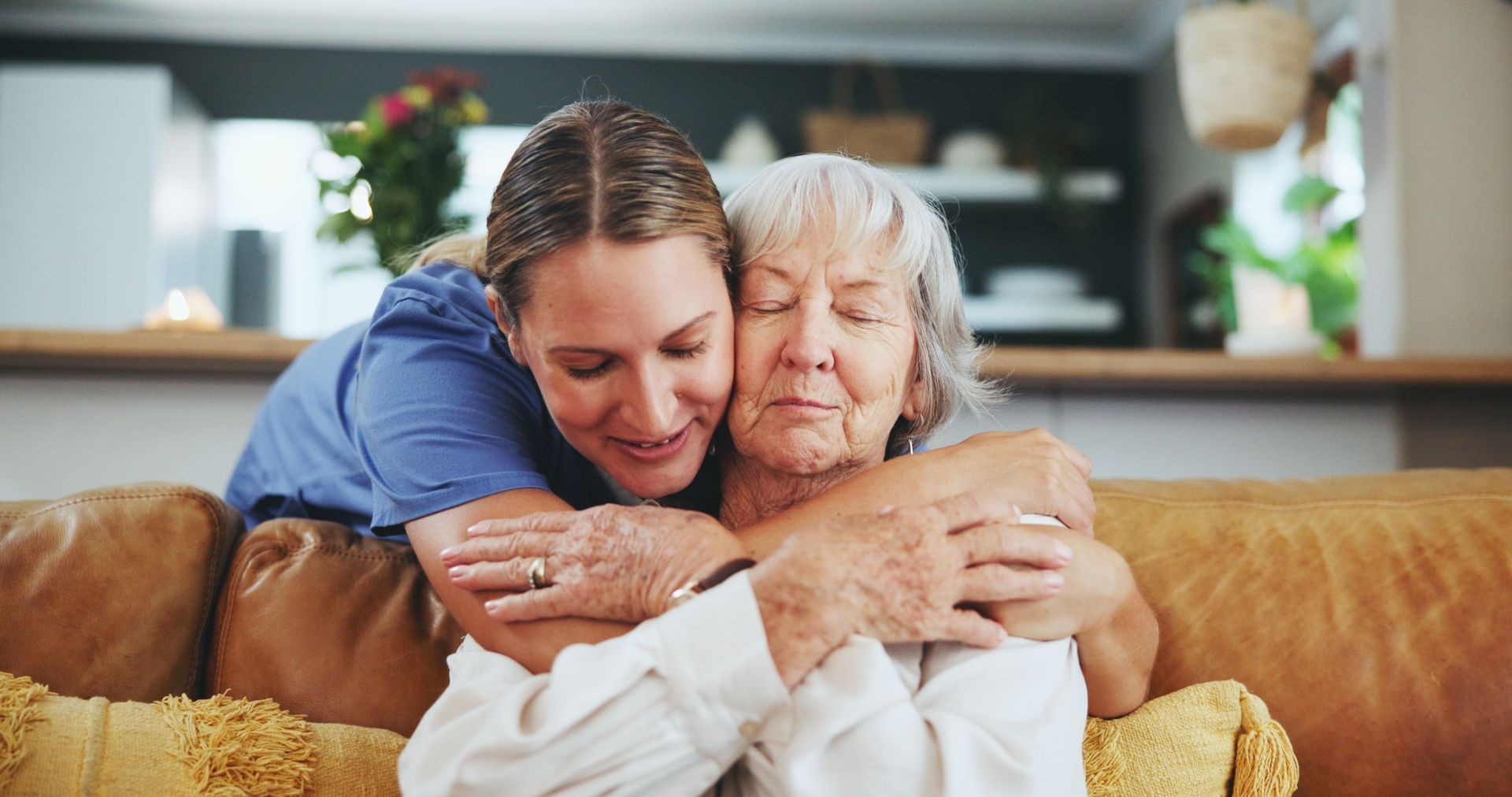 A caregiver hugs an elderly woman on a couch in a bright living room