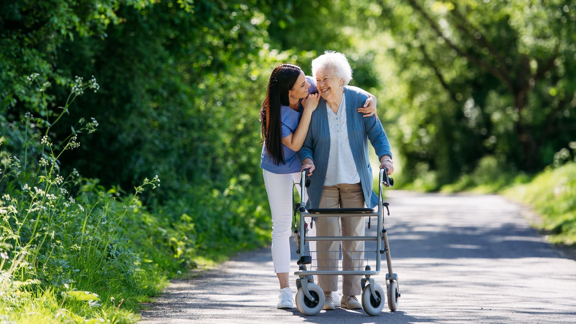 Older adult walking with a caregiver using a walker on a sunny tree-lined path