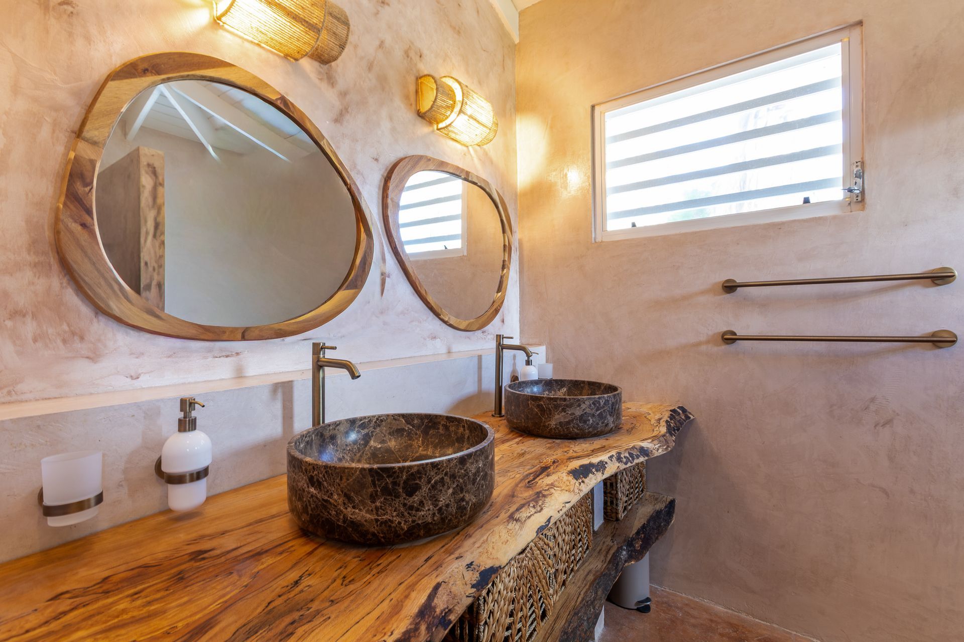 Badkamer met een houten badmeubel, twee stenen wastafels en organisch gevormde spiegels.