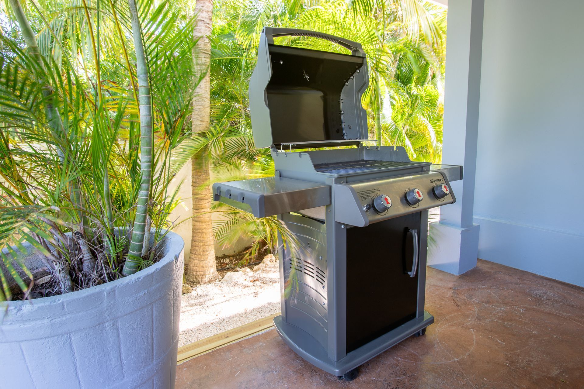 Grijze barbecue met open deksel, naast een potplant en een gebouw.