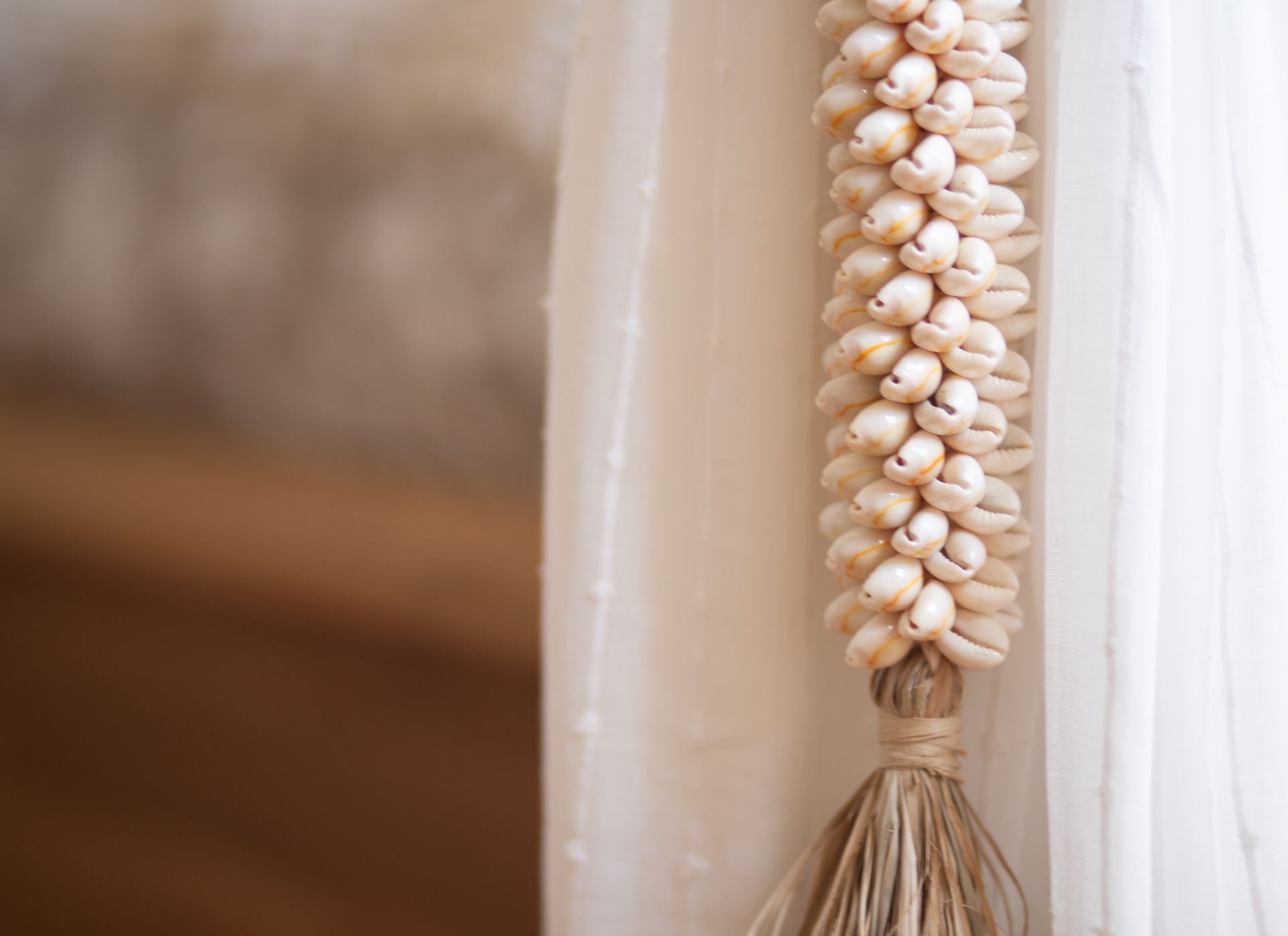 Een snoer schelpen hangt aan een doorschijnend wit gordijn, vastgebonden met touw en raffia.