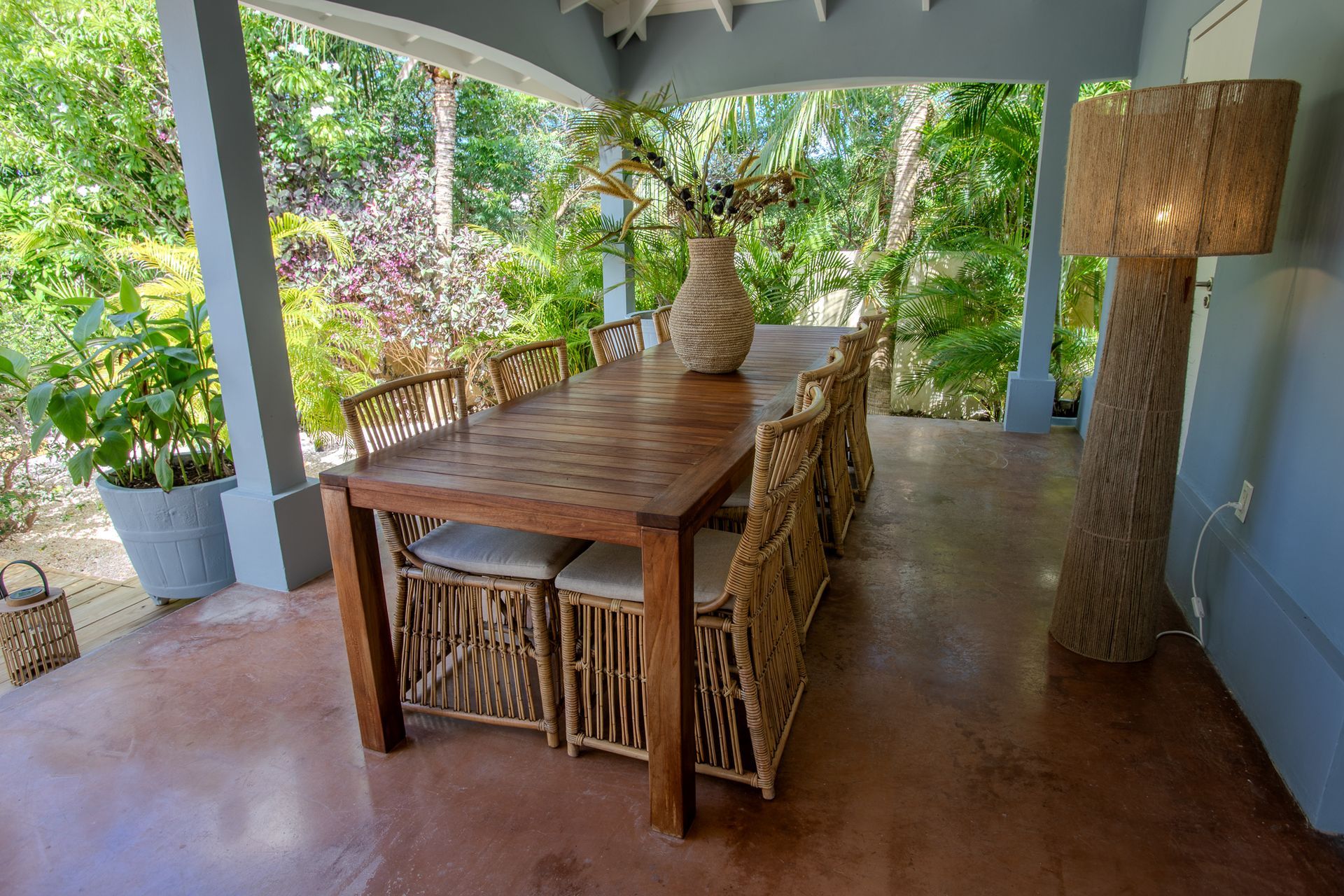 Overdekt terras met houten tafel, stoelen en staande lamp. Omgeven door weelderig groen.