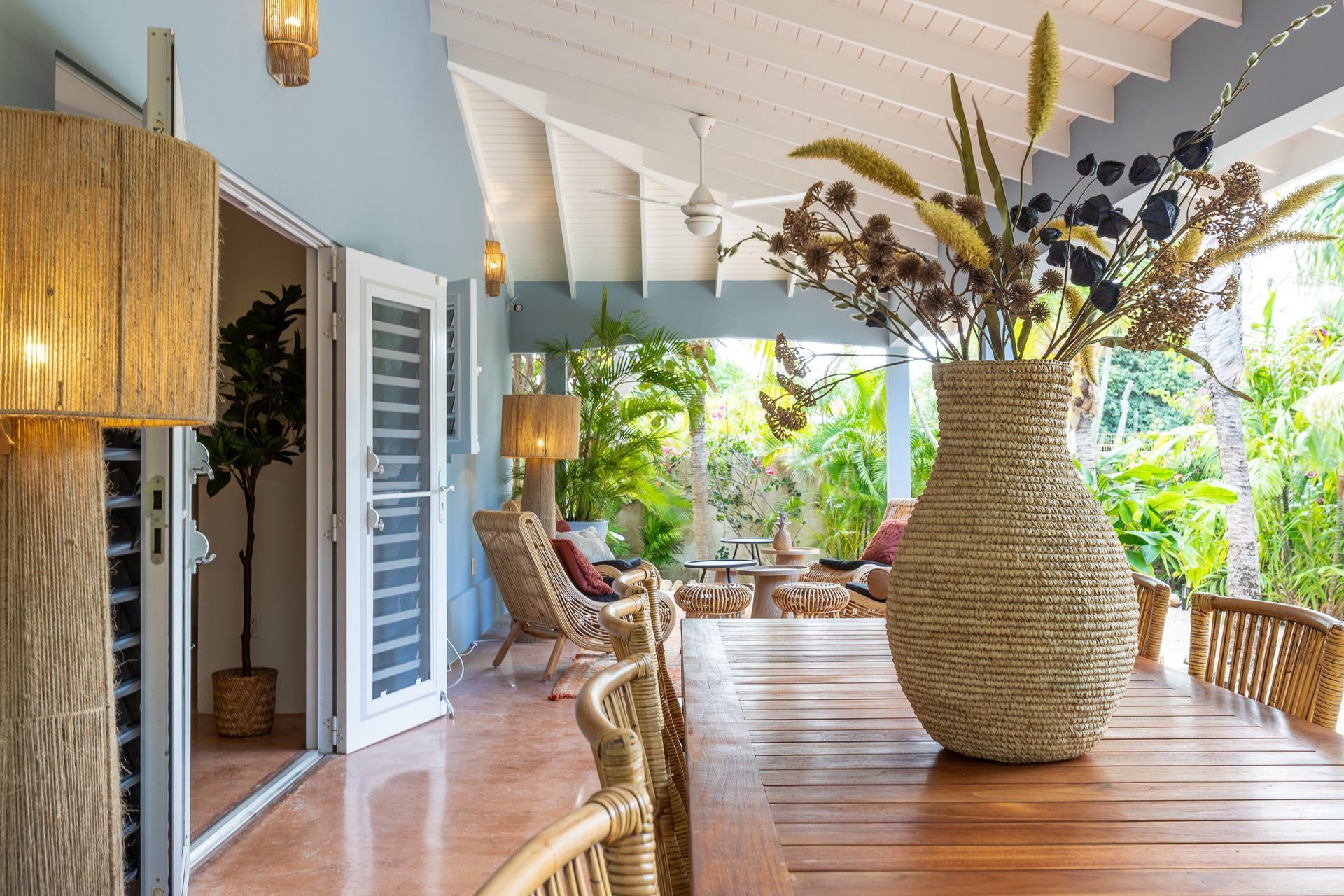 Overdekte veranda met eettafel, vaas met gedroogde bloemen, rotan stoelen en uitzicht op weelderig groen.