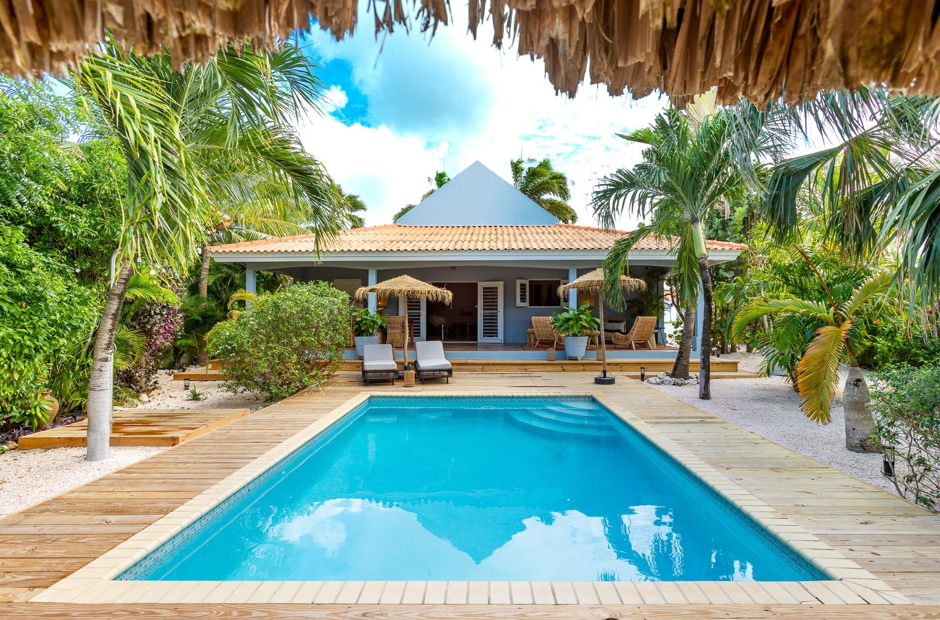 Zwembad met ligstoelen voor een huis, omgeven door weelderige tropische beplanting.
