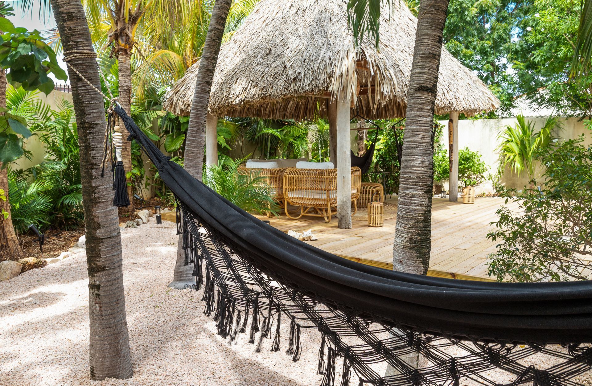 Een zwarte hangmat hangt tussen de bomen in een tropische buitenruimte, met op de achtergrond een eethoek met een rieten dak.