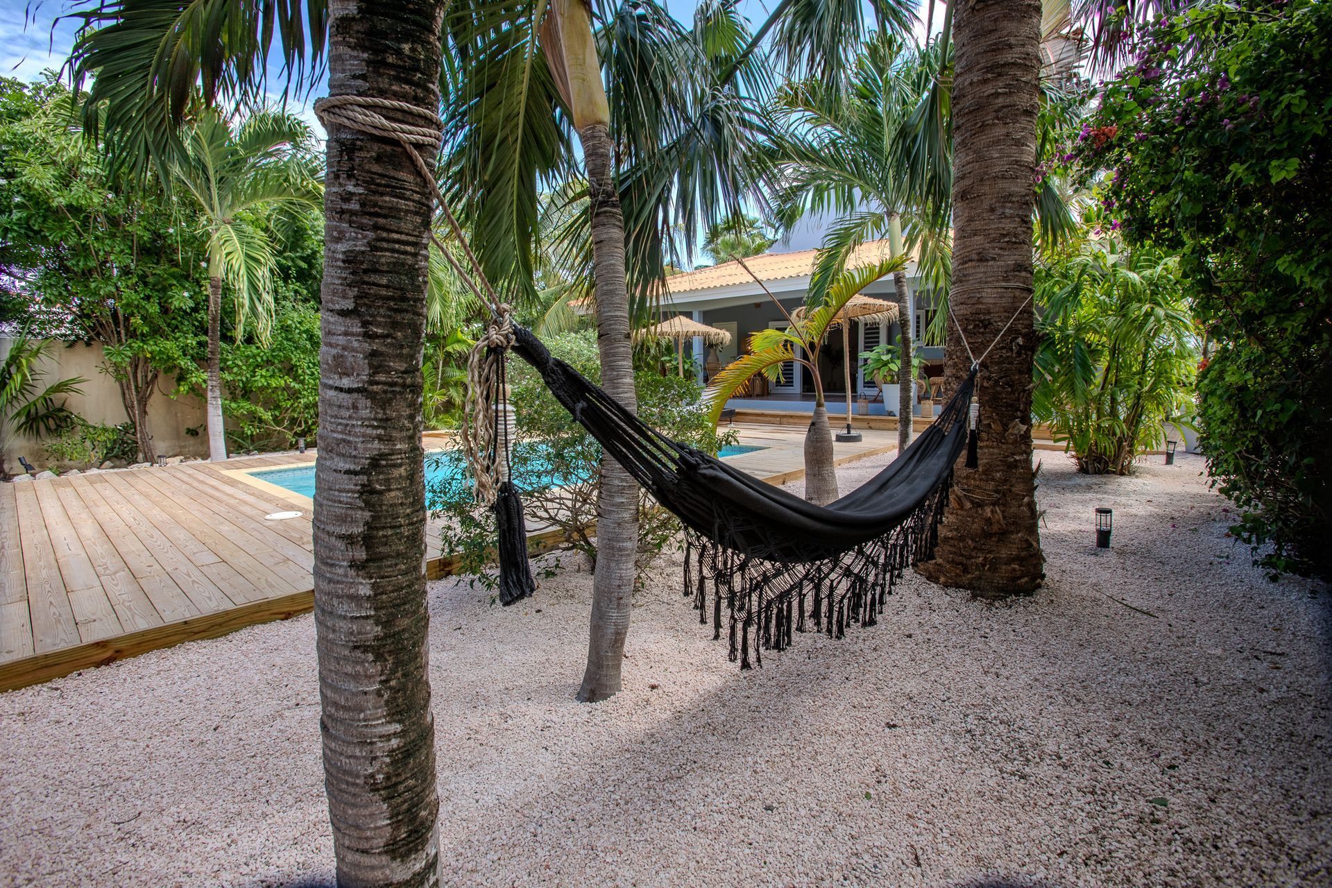 Een zwarte hangmat hangt tussen palmbomen in een tuin met een witte stenen ondergrond, een zwembad en een huis.