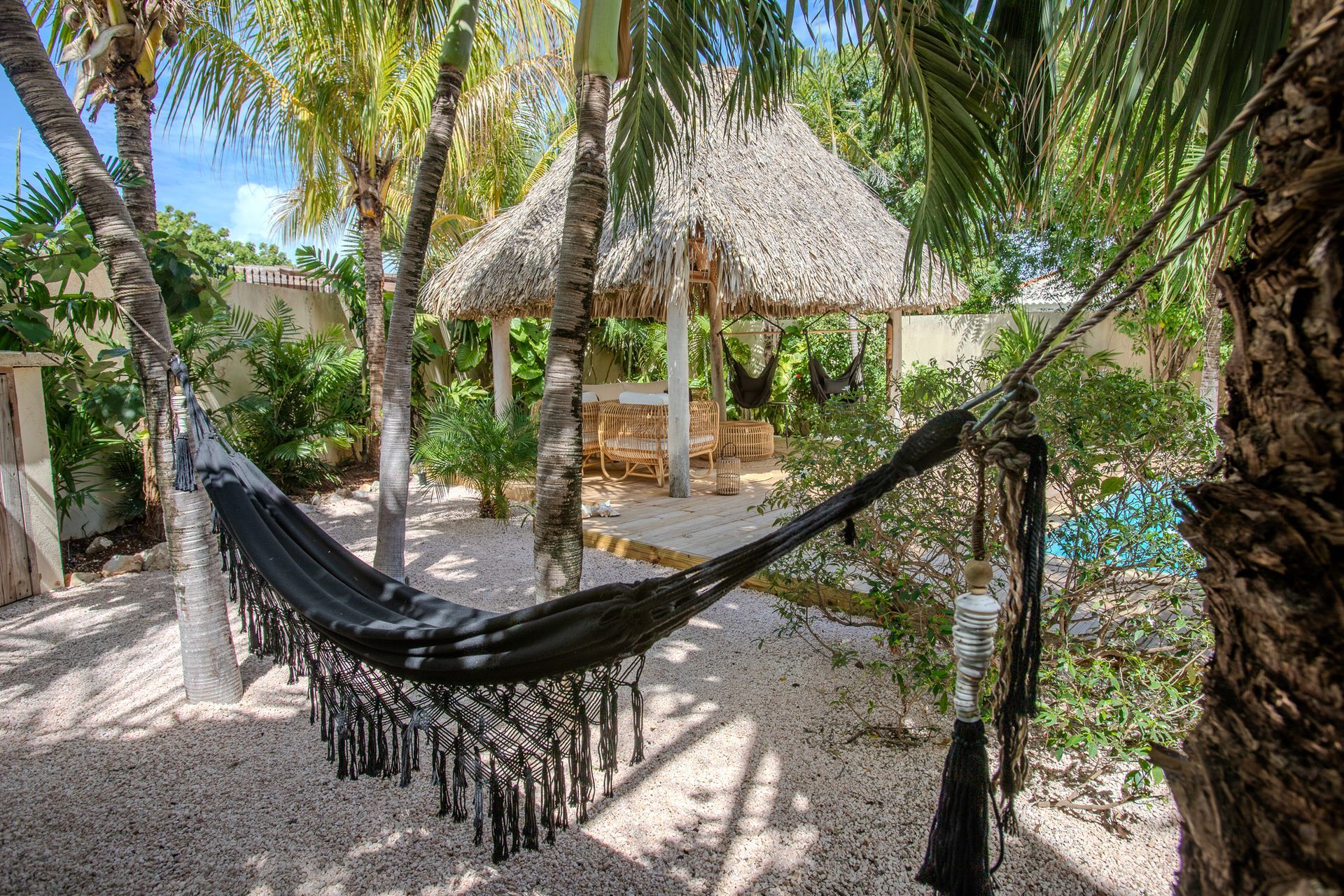 Een zwarte hangmat hangt tussen de bomen in een tropische achtertuin met een prieel met rieten dak.