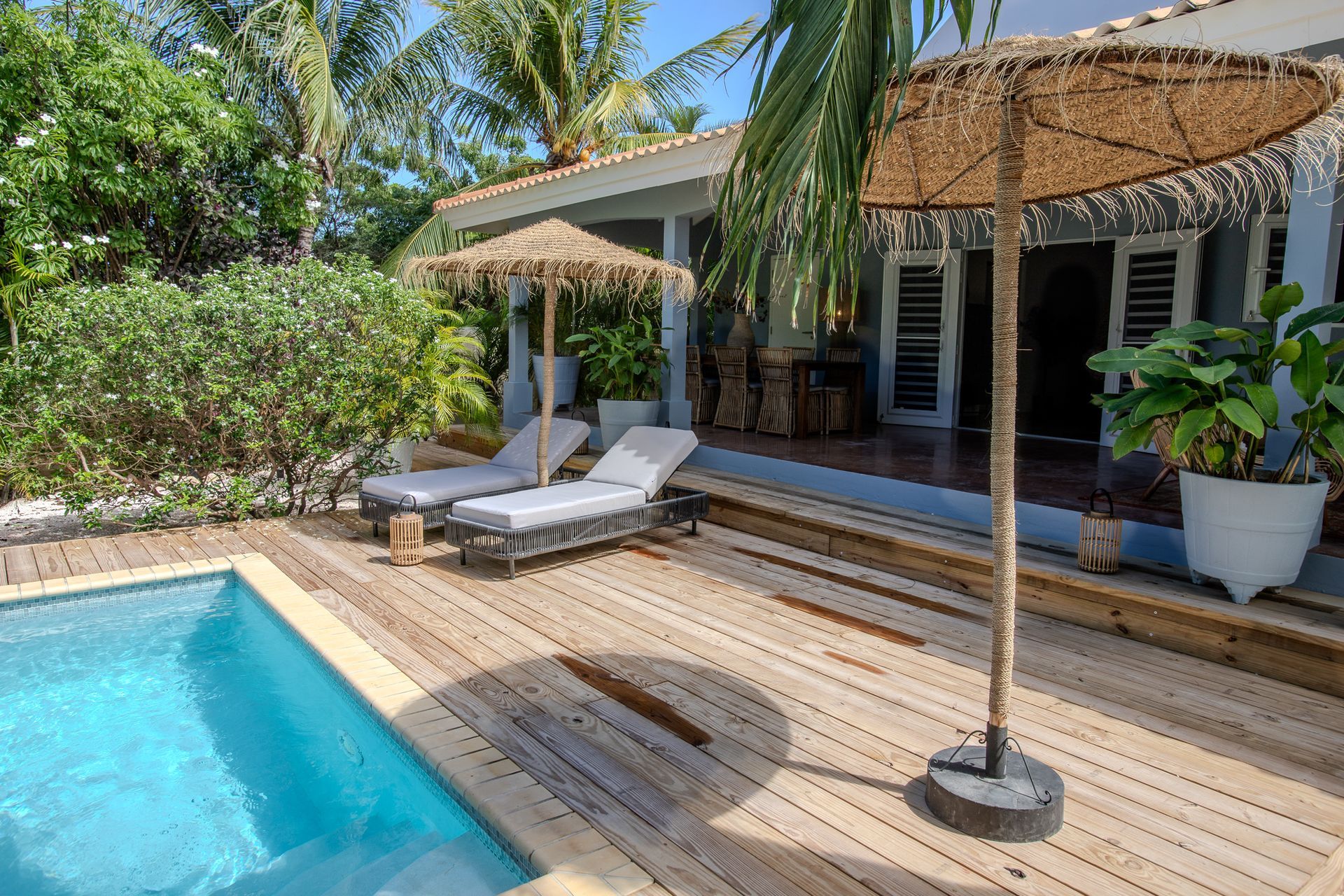 Een tafereel bij het zwembad: een houten terras, ligstoelen, rieten parasols en een huis met een veranda.