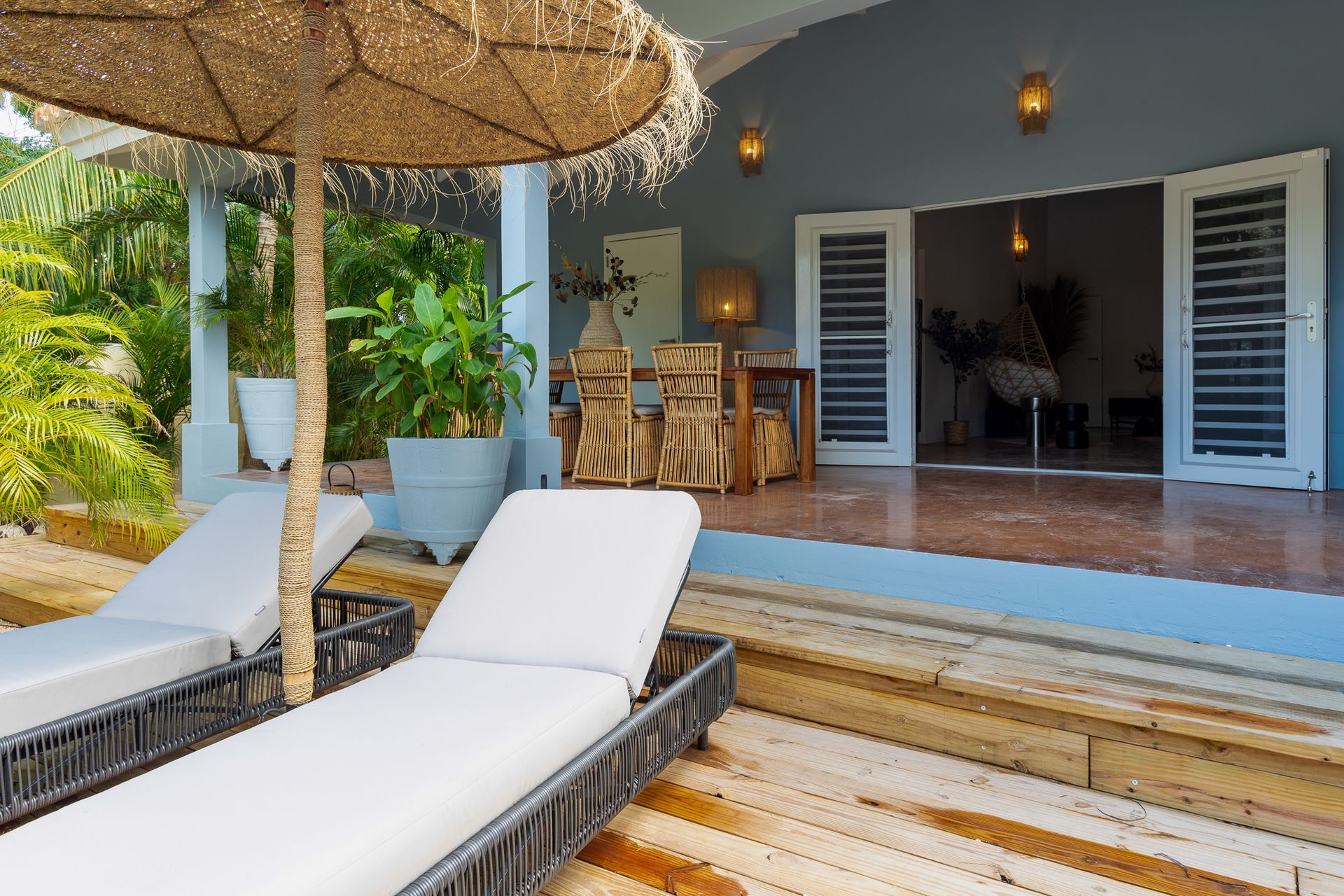 Twee ligstoelen onder een rieten parasol op een houten terras, dat uitkomt op een patio met een eethoek.
