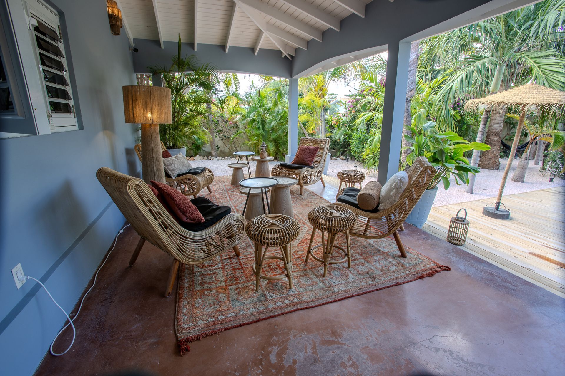 Overdekt terras met rieten meubels en een vloerkleed met patroon, met uitzicht op een tropische tuin.