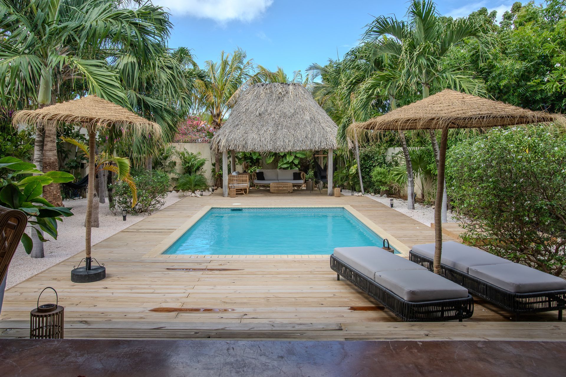 Zwembad met rieten parasols, ligstoelen en een prieel, omgeven door weelderig groen onder een blauwe hemel.