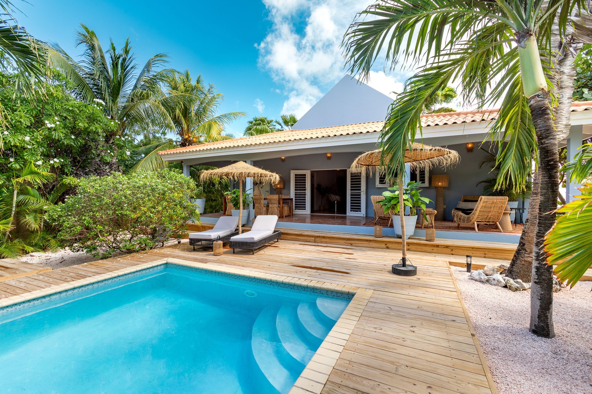 Vakantiehuis met zwembad en tropische sfeer, ligstoelen, palmbomen en een helderblauwe hemel.