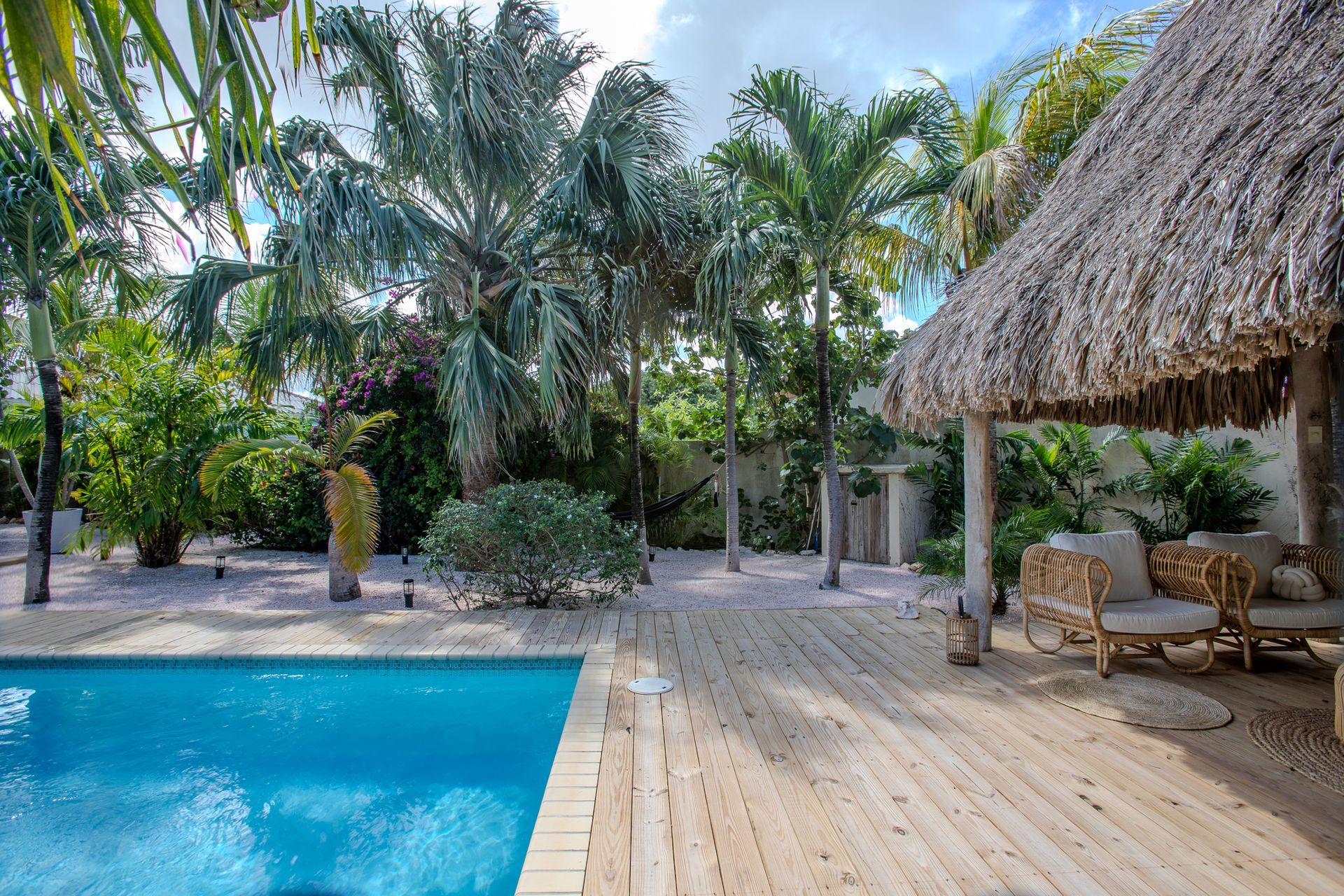 Zwembadgedeelte met blauw water, houten terras, cabana met rieten dak en tropische begroeiing.
