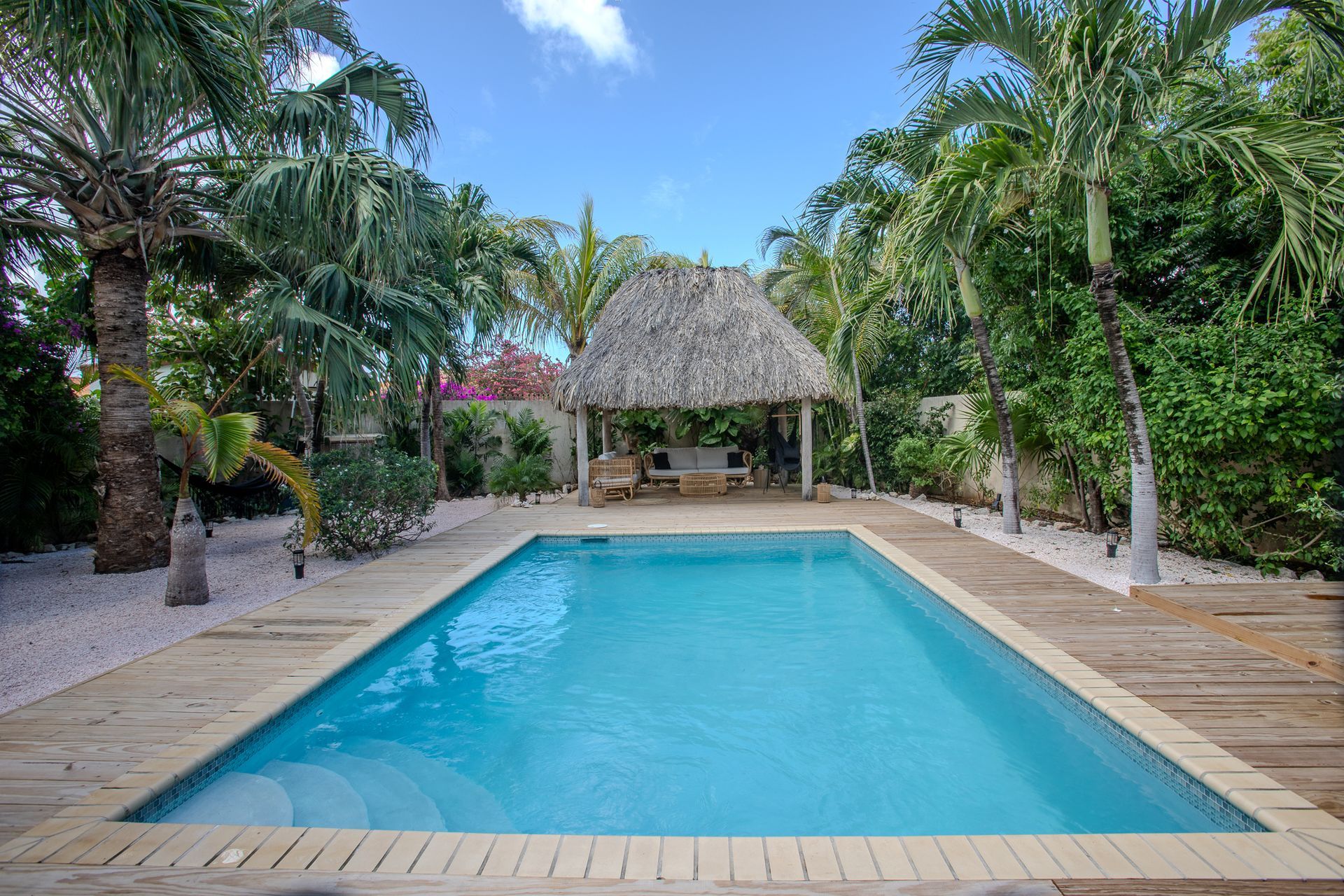 Zwembad in een tropische omgeving met een cabana met rieten dak, blauw water en palmbomen.