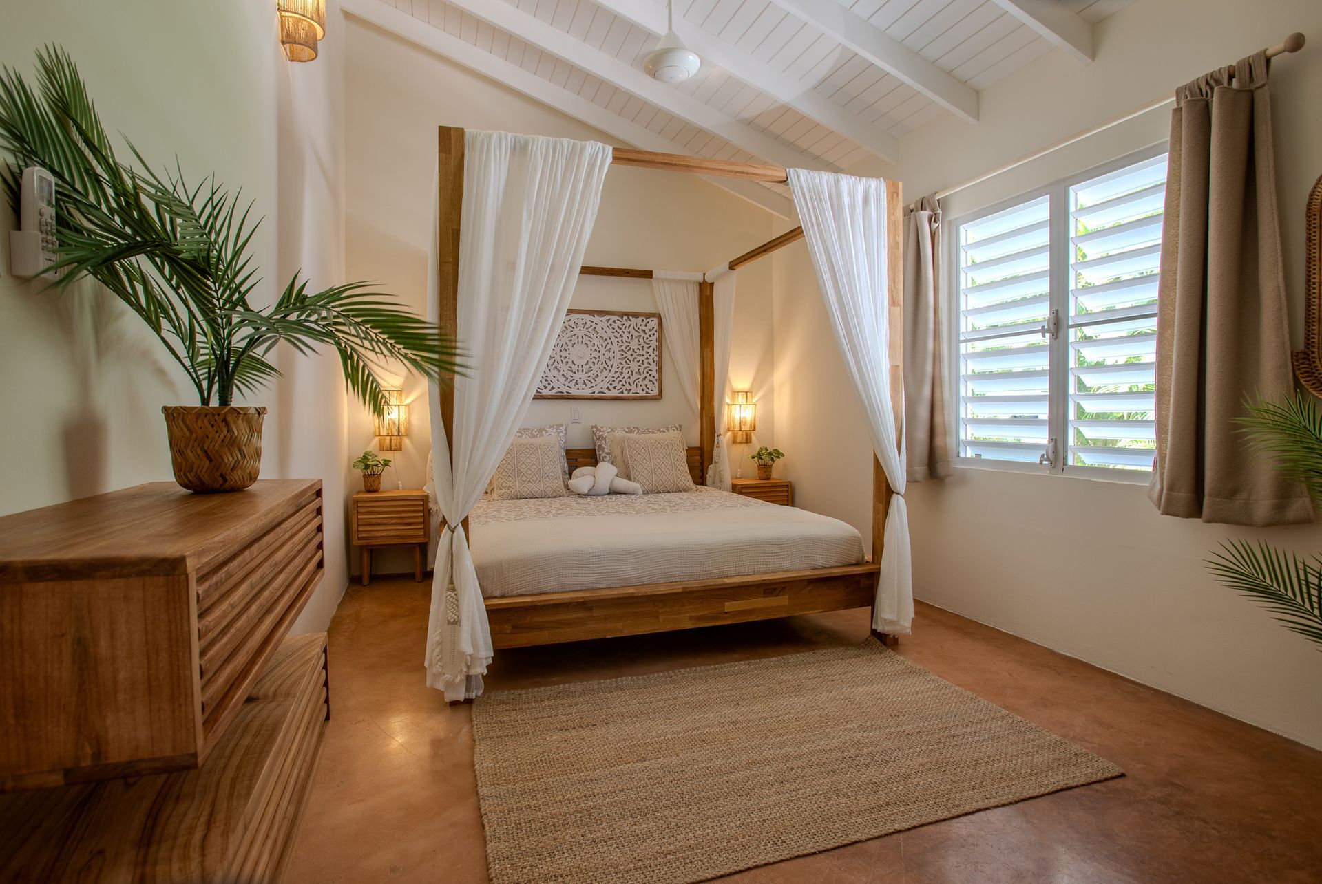 Slaapkamer met houten hemelbed, transparante gordijnen, meubels van natuurlijk hout, vloerkleed, raam met luiken en planten.