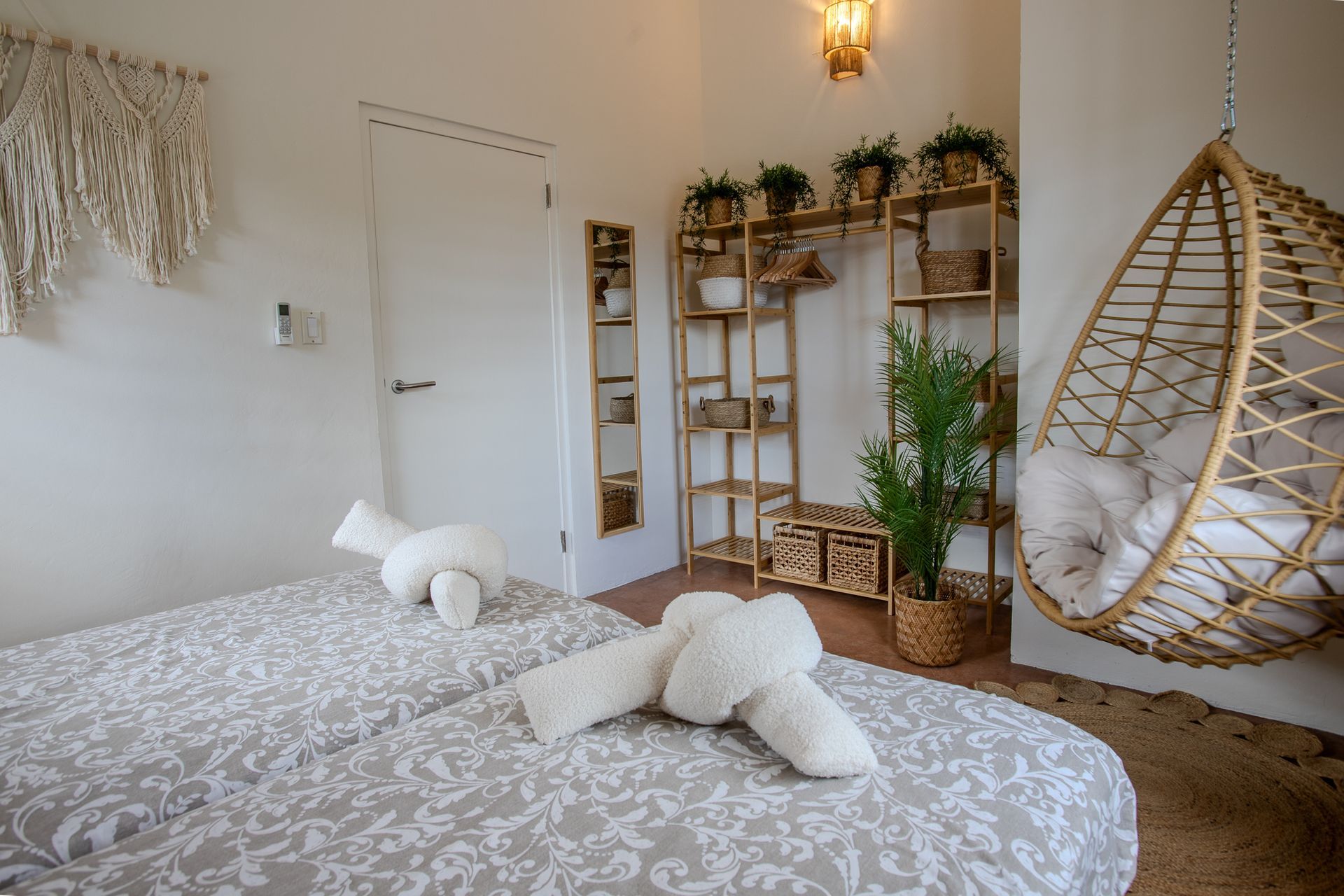 Slaapkamer met twee eenpersoonsbedden, een hangstoel, houten planken met planten en een geweven macramé wandkleed.