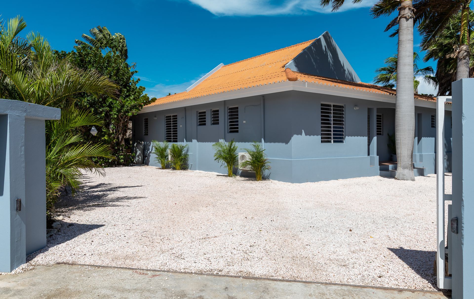 Grijs huis met oranje dak, witte grindtuin, blauwe lucht.