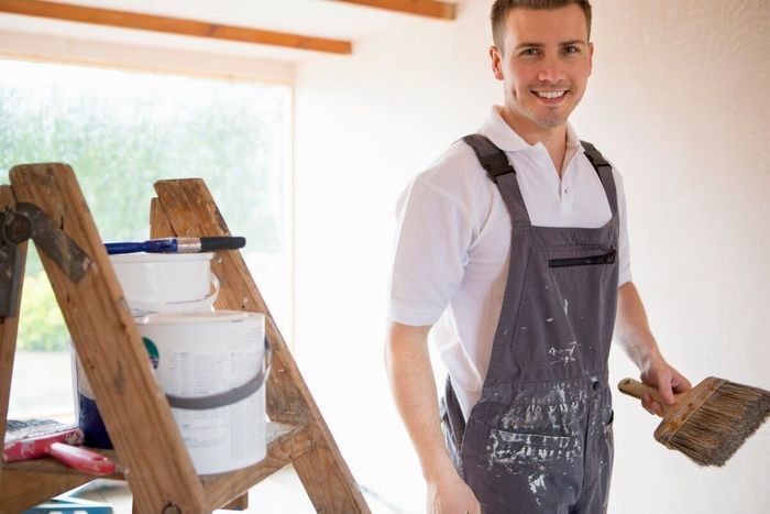 Sunderland painter smiling, holding brush, standing near ladder with eco-friendly paint cans. Light-filled room, partially painted walls by Foxglove Decor.