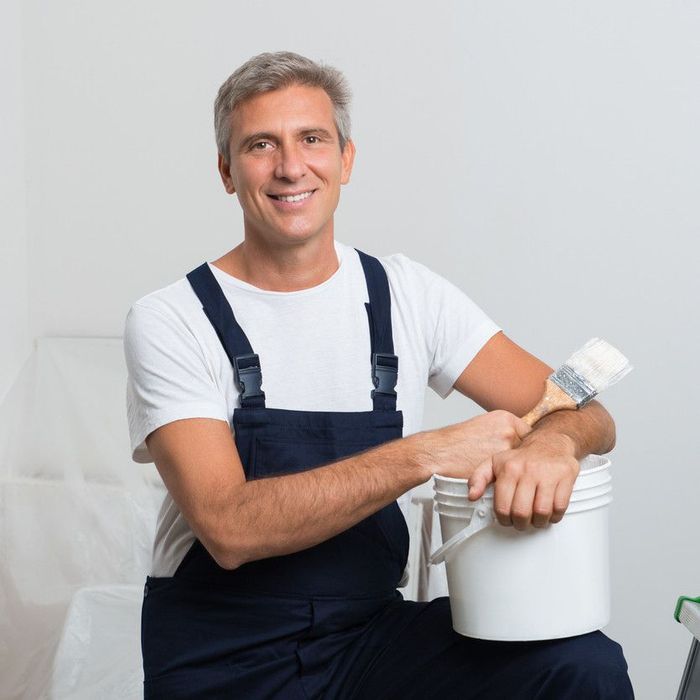 Sunderland painter smiles, holding brush and paint bucket, wearing overalls, in a room.